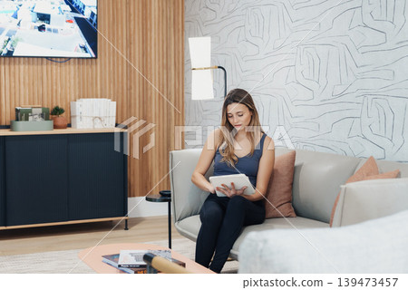 Female Engaged With Tablet On Sofa. Woman Deeply Involved In Electronic Notetaking At Home. Lady Absorbed In Educational Activities Using Her Tablet While Seated In Cozy Setting 139473457