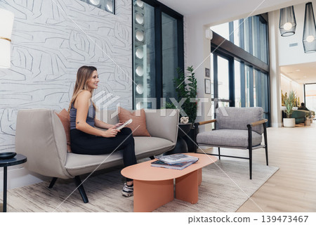 White Woman Entrepreneur Sitting On Sofa Reviewing Tablet In Bright Modern Coworking Lounge With Floortoceiling Windows, Indoor Plants, Neutral Furnishings, Relaxed Posture, Waiting Area White Woman Entrepreneur Sitting On Sofa Reviewing Tablet In Bright Modern Coworking Lounge With Floortoceiling Windows, Indoor Plants, Neutral Furnishings, Relaxed Posture, Waiting Area 139473467