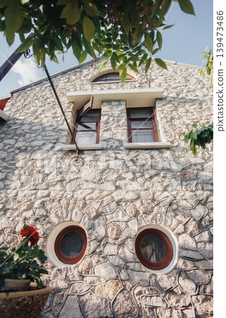 Sunlit Stone Facade With Round Windows, Arched Upper Window Framed In Wood, Terracotta Planter Brimming With Geranium, Climbing Vines And Leafy Canopy Casting Dappled Shadows, Textured 139473486
