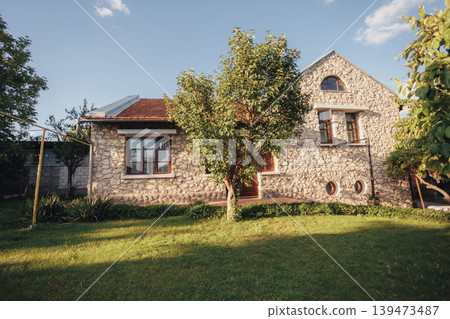 Stone House Exterior With Sunlit Garden, Writers Countryside Retreat Bathed In Warm Afternoon Light Stone Facade, Tiled Roof, Wooden Window Frames, Manicured Lawn, Mature Tree Casting Stone House Exterior With Sunlit Garden, Writers Countryside Retreat Bathed In Warm Afternoon Light Stone Facade, Tiled Roof, Wooden Window Frames, Manicured Lawn, Mature Tree Casting 139473487