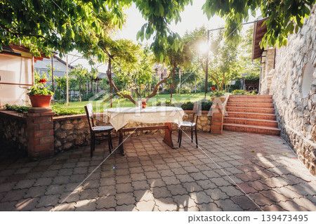 Sunlit Patio Table Under Leafy Trees, Stone Floor, Potted Plants, Wrought Iron Chairs Creating Cozy Alfresco Dining Atmosphere Ideal For Afternoon Coffee, Intimate Gathering, Slowliving Sunlit Patio Table Under Leafy Trees, Stone Floor, Potted Plants, Wrought Iron Chairs Creating Cozy Alfresco Dining Atmosphere Ideal For Afternoon Coffee, Intimate Gathering, Slowliving 139473495