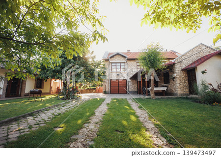 Spacious Stone Courtyard Framed By Trees And Trimmed Lawn, Dual Grass Tracks Leading To Wooden Garage Doors, Calm Midday Light, Vintage Charm And Tidy Landscape Suited For Real Estate Spacious Stone Courtyard Framed By Trees And Trimmed Lawn, Dual Grass Tracks Leading To Wooden Garage Doors, Calm Midday Light, Vintage Charm And Tidy Landscape Suited For Real Estate 139473497