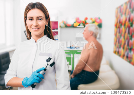 Doctor dermatologist with dermatoscope, smiling while looking at camera. Behind her sitting senior patient. Doctor dermatologist with dermatoscope, smiling while looking at camera. Behind her sitting senior patient. 139473520