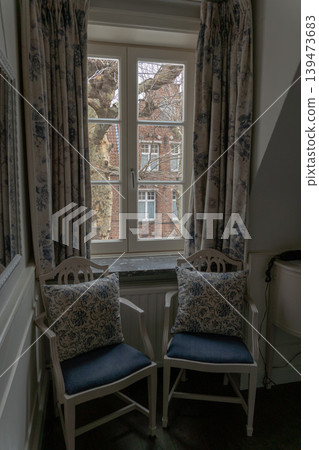 Cozy hotel room corner with upholstered chairs, patterned cushions and tall window with curtains. Hospitality interior, travel accommodation, classic decor, relaxing atmosphere, lifestyle Cozy hotel room corner with upholstered chairs, patterned cushions and tall window with curtains. Hospitality interior, travel accommodation, classic decor, relaxing atmosphere, lifestyle 139473683