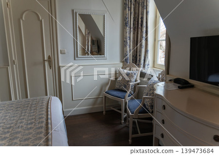 Elegant hotel room interior with upholstered chairs, mirror, dresser and window with patterned curtains. Hospitality design, cozy accommodation, classic decor, travel lifestyle, interior inspiration Elegant hotel room interior with upholstered chairs, mirror, dresser and window with patterned curtains. Hospitality design, cozy accommodation, classic decor, travel lifestyle, interior inspiration 139473684