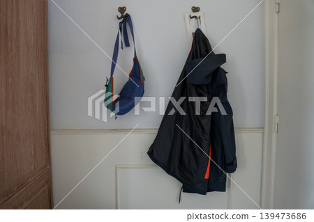 Wall hooks with hanging jacket, blue shoulder bag on white interior wall in home or hotel setting. Everyday organization, clothing storage, casual lifestyle, interior detail, accommodation concept Wall hooks with hanging jacket, blue shoulder bag on white interior wall in home or hotel setting. Everyday organization, clothing storage, casual lifestyle, interior detail, accommodation concept 139473686
