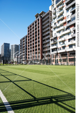 Urban football field beside tall residential buildings under clear sky. Urban football field beside tall residential buildings under clear sky. 139474027