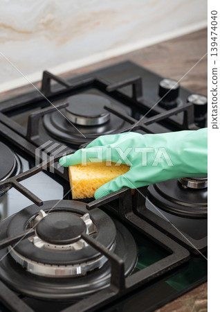 A close-up of a person's hand in a rubber glove cleaning a gas cooktop with a sponge. 139474040