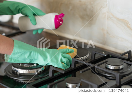 A close-up of a person's hand in a rubber glove cleaning a gas cooktop with a sponge. 139474041