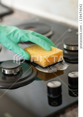 A close-up of a person's hand in a rubber glove cleaning a gas cooktop with a sponge. A close-up of a person's hand in a rubber glove cleaning a gas cooktop with a sponge. 139474042