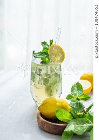 A summer drink with lemon, mint, and ice in a glass against a sunny window background. A summer drink with lemon, mint, and ice in a glass against a sunny window background. 139474053