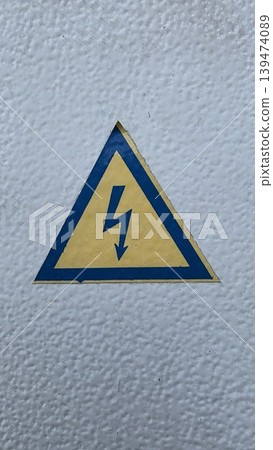 Yellow high voltage warning sign with a lightning bolt on a textured white metal electrical box surface. Concept of industrial safety and electrical hazard Yellow high voltage warning sign with a lightning bolt on a textured white metal electrical box surface. Concept of industrial safety and electrical hazard 139474089