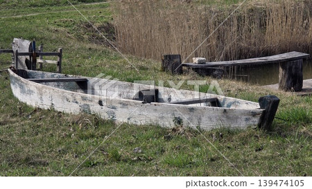 Weathered wooden boat on grassy pond shore near reeds and small pier, quiet rural scene for countryside, heritage, travel and nature concepts. Weathered wooden boat on grassy pond shore near reeds and small pier, quiet rural scene for countryside, heritage, travel and nature concepts. 139474105