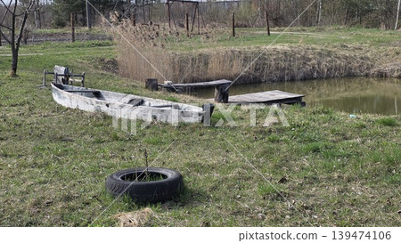 Weathered old wooden boat and a tire lying on green grass near a small pond with a wooden pier in the countryside. Concept of rural life and solitude Weathered old wooden boat and a tire lying on green grass near a small pond with a wooden pier in the countryside. Concept of rural life and solitude 139474106
