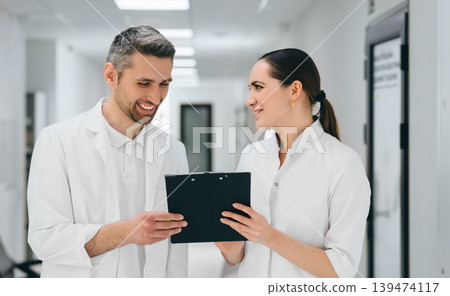 Two doctors with clipboard communicate standing at hospital corridor. Doctors laughing and having friendly emotions Two doctors with clipboard communicate standing at hospital corridor. Doctors laughing and having friendly emotions 139474117