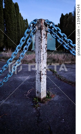 Old peeling white metal post with blue chains on cracked asphalt against row of cypress trees and turquoise building at twilight. Gloomy atmosphere of an abandoned territory Old peeling white metal post with blue chains on cracked asphalt against row of cypress trees and turquoise building at twilight. Gloomy atmosphere of an abandoned territory 139474144