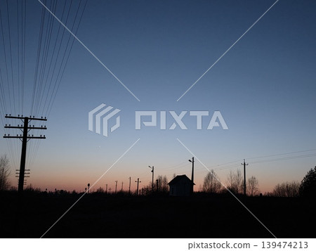 Silhouette of a small rural house and multiple power lines against a clear pink and blue sunset sky. Concept of remote life and energy supply 139474213