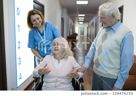 senior woman in wheelchair talking with nurse in a hospital behind her standing senior husband. senior woman in wheelchair talking with nurse in a hospital behind her standing senior husband. 139474250
