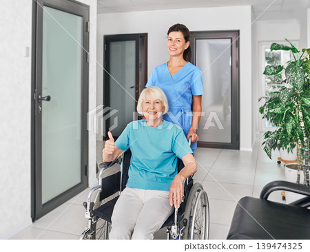 Friendly geriatric nurse assisting senior woman in a wheelchair at nursing home corridor. Medical care for people with disabilities Friendly geriatric nurse assisting senior woman in a wheelchair at nursing home corridor. Medical care for people with disabilities 139474325