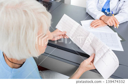 Elderly patient looking at the results of a cardiogram of her heart in a cardiologist's office. Diagnostics and treatment of arrhythmias, coronary heart disease and heart failure Elderly patient looking at the results of a cardiogram of her heart in a cardiologist's office. Diagnostics and treatment of arrhythmias, coronary heart disease and heart failure 139474332