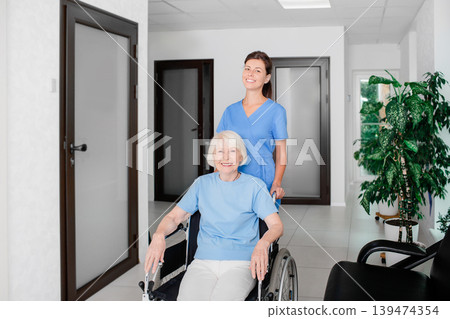 Friendly nurse taking care of the senior patient in a wheelchair at the hospital corridor Friendly nurse taking care of the senior patient in a wheelchair at the hospital corridor 139474354