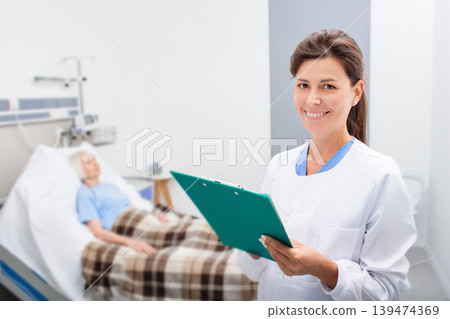 Smiling doctor is holding a medical record while standing in a hospital ward. Therapist is happy because the patient's tests are normal. In the background, an elderly woman in a hospital bed. Smiling doctor is holding a medical record while standing in a hospital ward. Therapist is happy because the patient's tests are normal. In the background, an elderly woman in a hospital bed. 139474369