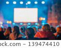 Rear view of blurred crowd watches large screen at night outdoor event with lights, bokeh, people silhouettes. Concept of screen advertising, cinema, festival, presentation, entertainment, copy space 139474410