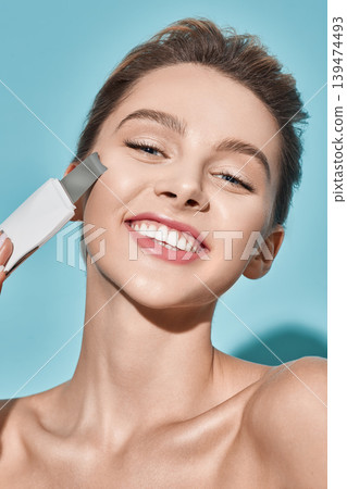 Happy woman with ultrasound scrubber treats her skin for remove blackheads and cleansing facial skin, on blue background. Ultrasonic facial peeling Happy woman with ultrasound scrubber treats her skin for remove blackheads and cleansing facial skin, on blue background. Ultrasonic facial peeling 139474493