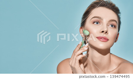 Beautiful woman using stone facial roller for elastic perfect skin, isolated on blue background. Jade roller with natural quartz stones at cosmetology 139474495