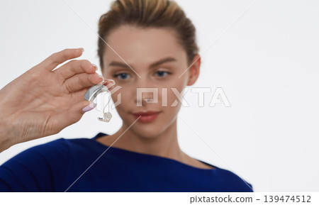 Treatment of deafness in adult people, hearing solutions. Beautiful woman holding latest generation hearing aid in her hand on foreground Treatment of deafness in adult people, hearing solutions. Beautiful woman holding latest generation hearing aid in her hand on foreground 139474512