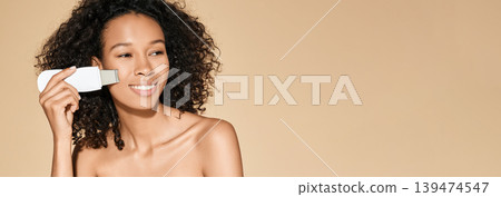 Curly African American woman taking care of her skin with ultrasound scrubber, removing acne and cleansing skin of her face 139474547