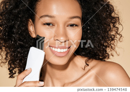 African American woman with ultrasound scrubber treating her skin for remove blackheads and cleansing facial skin, on beige background. Ultrasonic facial peeling 139474548