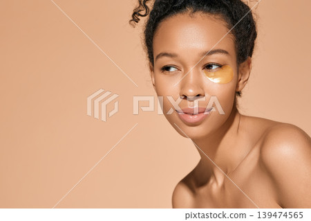 African American woman with golden hydrogel patches under her eyes to relieve puffiness during cosmetic procedures, isolated on beige background. Skin care 139474565