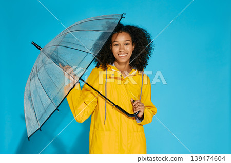 Young African American woman advertises trendy yellow raincoat and transparent umbrella for autumn season standing in studio on blue background Young African American woman advertises trendy yellow raincoat and transparent umbrella for autumn season standing in studio on blue background 139474604