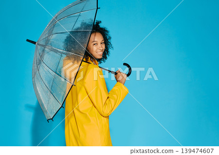 African American woman in yellow waterproof raincoat with transparent umbrella isolated on blue studio background. Autumn season concept 139474607