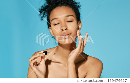 African American applies serum on her face with pipette, on blue background. Facial skin care with treatment cosmetic oil and serum African American applies serum on her face with pipette, on blue background. Facial skin care with treatment cosmetic oil and serum 139474617