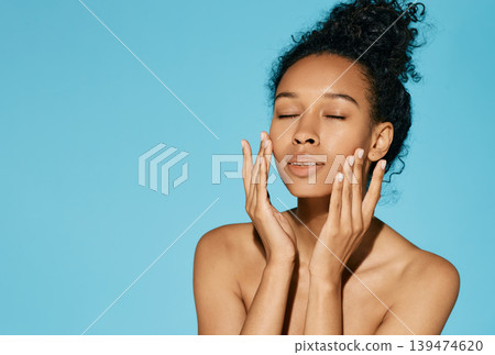 Clean skin concept. Curly sensual woman with smooth fresh face skin holding hands near her beauty face on blue background Clean skin concept. Curly sensual woman with smooth fresh face skin holding hands near her beauty face on blue background 139474620
