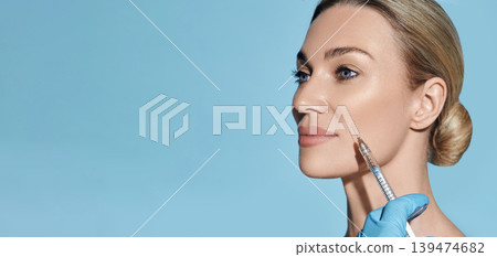 Beautician doing beauty injection into nasolabial folds on woman face with mimic wrinkles near mouth, on blue background. Botulinum toxin injections 139474682