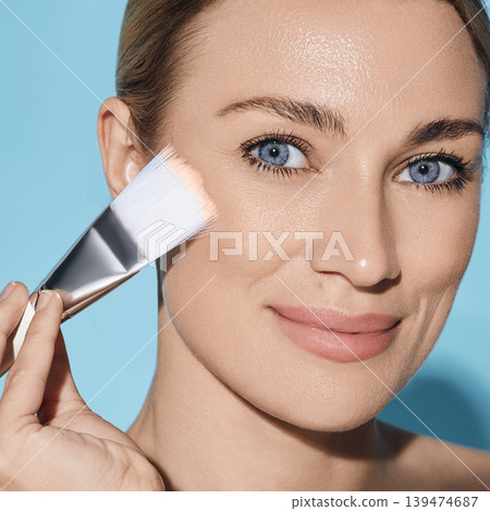 Beautiful woman applying tone cream on her perfect face with cosmetic brush, isolated on blue background. Facial cosmetics, foundation Beautiful woman applying tone cream on her perfect face with cosmetic brush, isolated on blue background. Facial cosmetics, foundation 139474687