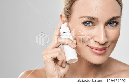 Positive caucasian woman applies moisturizing cream on her face, on grey background. Face cream moisturizing skin, skincare Positive caucasian woman applies moisturizing cream on her face, on grey background. Face cream moisturizing skin, skincare 139474692