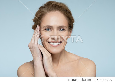 Perfect middle-aged female face. Beautiful woman with clean and healthy skin with hands near face, isolated on blue background Perfect middle-aged female face. Beautiful woman with clean and healthy skin with hands near face, isolated on blue background 139474709