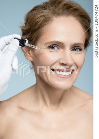 Facial skin care with treatment cosmetic oil and serum. Beautiful Caucasian woman applying serum on her face, isolated on blue background Facial skin care with treatment cosmetic oil and serum. Beautiful Caucasian woman applying serum on her face, isolated on blue background 139474724