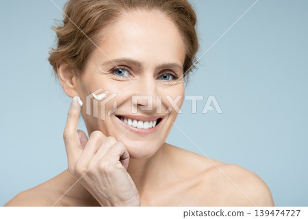 Middle-age woman applying cream and anti-aging cosmetics on face, on blue background. Skin care for women with anti-aging cosmetics Middle-age woman applying cream and anti-aging cosmetics on face, on blue background. Skin care for women with anti-aging cosmetics 139474727