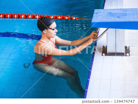 Young woman swimmer in a swimsuit getting ready to start swimming in a swimming pool. Professional swimmer near the starting block. Young woman swimmer in a swimsuit getting ready to start swimming in a swimming pool. Professional swimmer near the starting block. 139474747