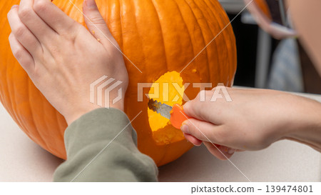 Person carving a pumpkin for Halloween with a small knife, showcasing the orange flesh and hollowed interior on a light-colored table 139474801