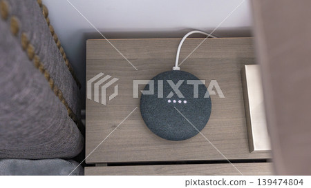 Smart home speaker on wooden nightstand with charging cable, surrounded by soft furnishings and a decorative box in a modern interior setting Smart home speaker on wooden nightstand with charging cable, surrounded by soft furnishings and a decorative box in a modern interior setting 139474804
