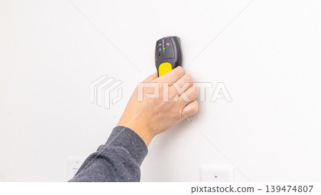 Mans hand holding yellow stud finder against white wall near electrical outlet after installation. Home improvement, checking placement, DIY verification, tool usage. Mans hand holding yellow stud finder against white wall near electrical outlet after installation. Home improvement, checking placement, DIY verification, tool usage. 139474807