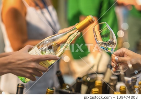 White wine tasting at indoor event with bottle and glass close-up. Wine being poured from glass bottle into tasting glass, blurred people, bottles, warm lights, shallow depth of field in background White wine tasting at indoor event with bottle and glass close-up. Wine being poured from glass bottle into tasting glass, blurred people, bottles, warm lights, shallow depth of field in background 139474808