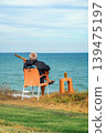 Elderly musician sits on chair by sea, playing guitar near coast with wine drink, creating travel, lifestyle, retirement, vacation themes. Concept of leisure, solitude, freedom with music by ocean 139475197