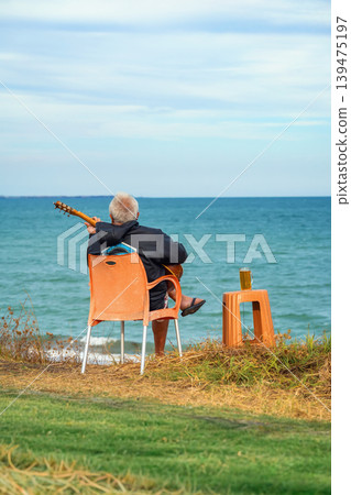 Elderly musician sits on chair by sea, playing guitar near coast with wine drink, creating travel, lifestyle, retirement, vacation themes. Concept of leisure, solitude, freedom with music by ocean Elderly musician sits on chair by sea, playing guitar near coast with wine drink, creating travel, lifestyle, retirement, vacation themes. Concept of leisure, solitude, freedom with music by ocean 139475197